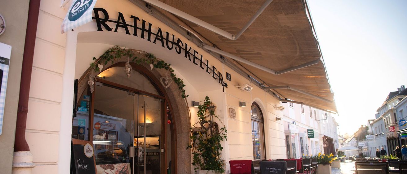 Außenansicht des Rathauskeller Restaurants mit einem Vordach, einer Tafel mit Speisekarte und Tischen und Stühlen draußen. Eine Flagge hängt über dem Eingang.