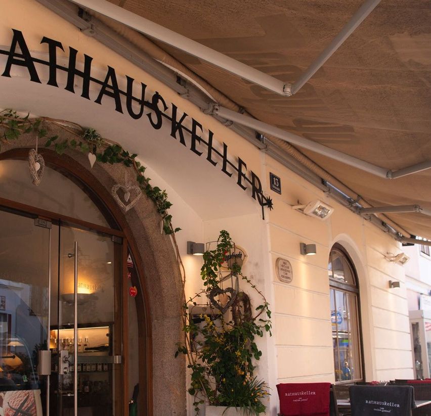 Außenansicht des Rathauskeller Restaurants mit einem Vordach, einer Tafel mit Speisekarte und Tischen und Stühlen draußen. Eine Flagge hängt über dem Eingang.