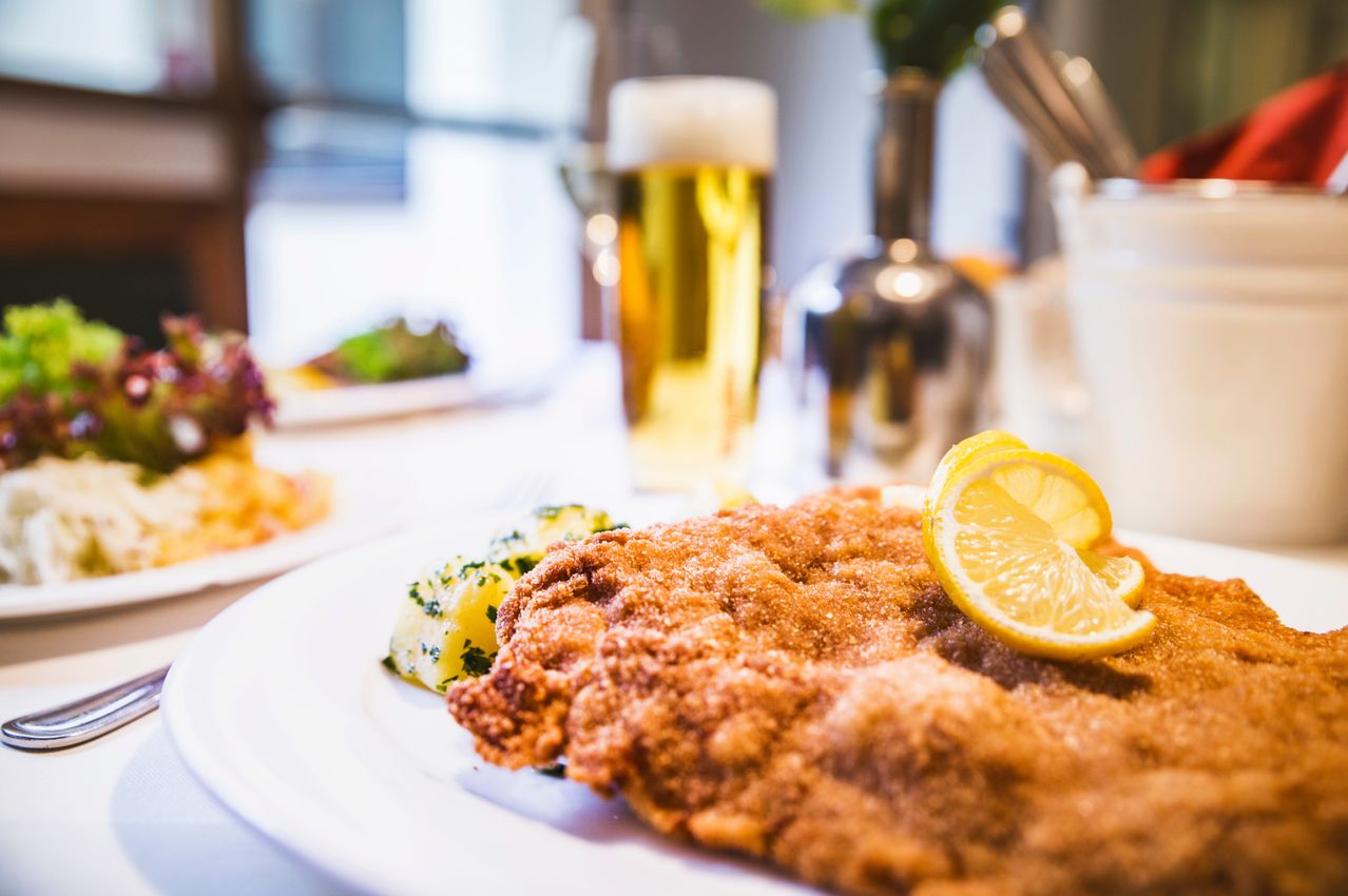 Nahaufnahme eines traditionellen deutschen Gerichts, Schnitzel, serviert mit einer Beilage Kartoffelpüree und einer Zitronenscheibe, mit einem Glas Bier im Hintergrund.