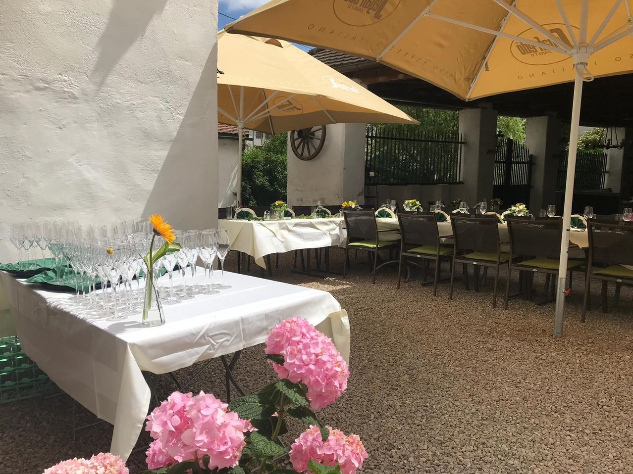 Der Außenbereich eines Restaurants mit Tischen und Stühlen unter Schirmen, geschmückt mit weißen Tischdecken und Blumen. Ein Tisch davor ist mit Weingläsern gedeckt.