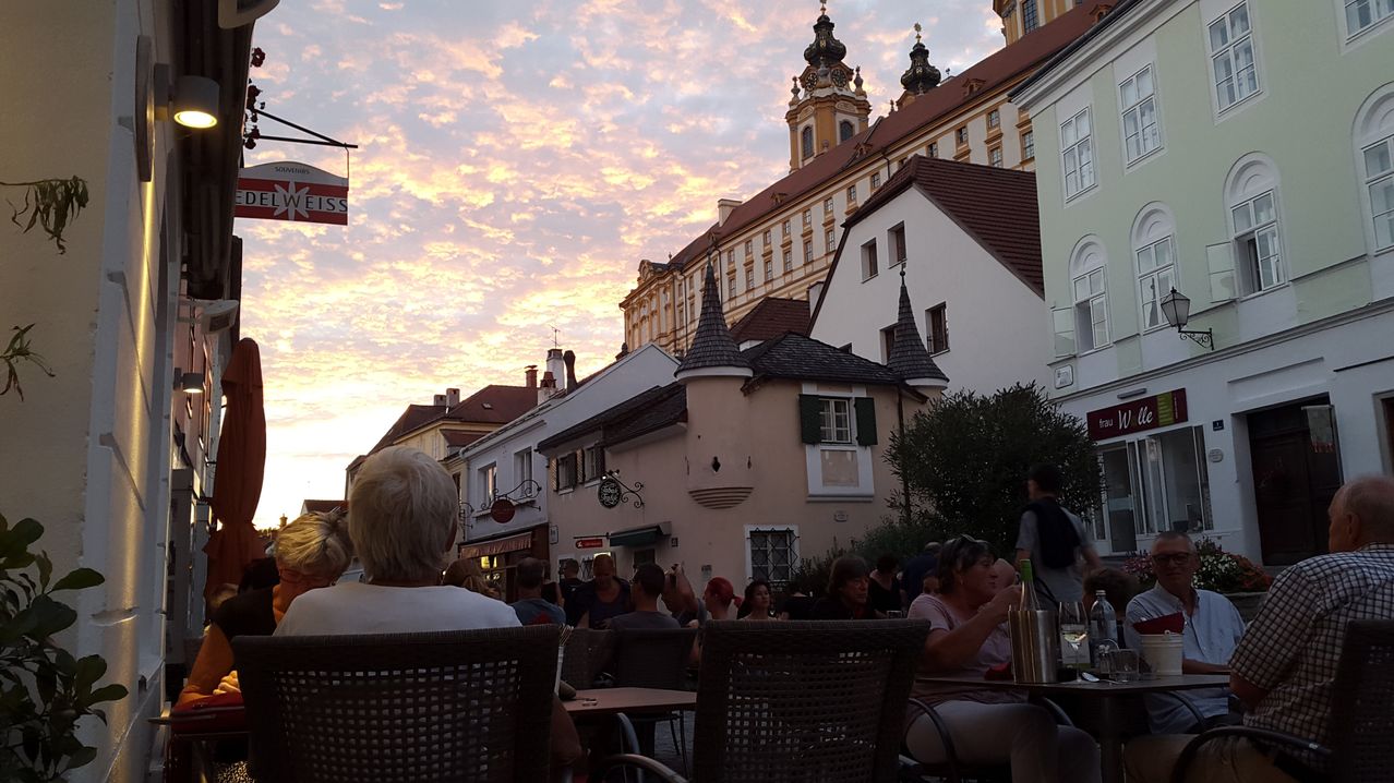 Ein Sonnenuntergang über einem europäischen Stadtplatz. Menschen sitzen an Tischen vor einem Gebäude mit Glockenturm. Der Himmel ist orange und rosa.