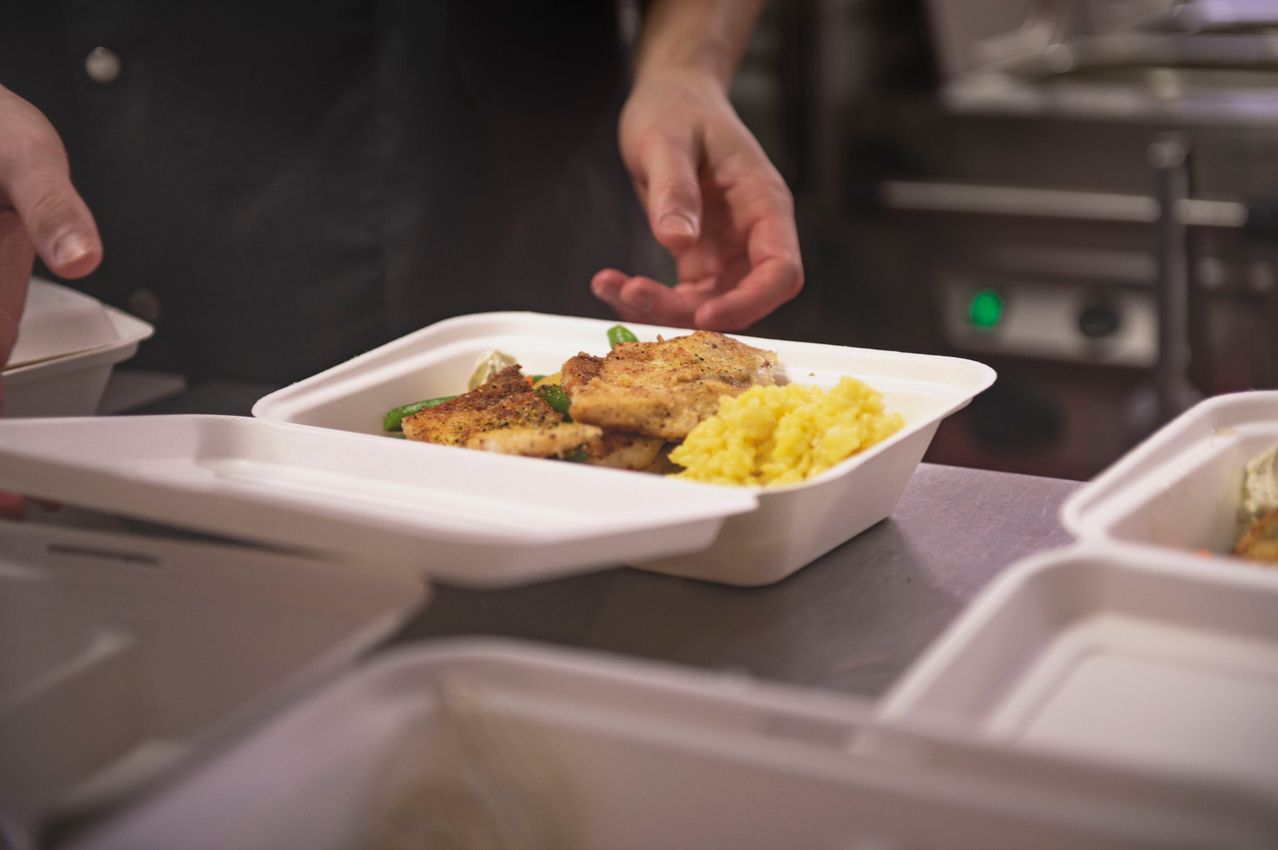 Eine Nahaufnahme einer Person, die einen weißen Styroporbehälter mit gebratenem Hühnchen, grünen Bohnen und gelbem Reis würzt.