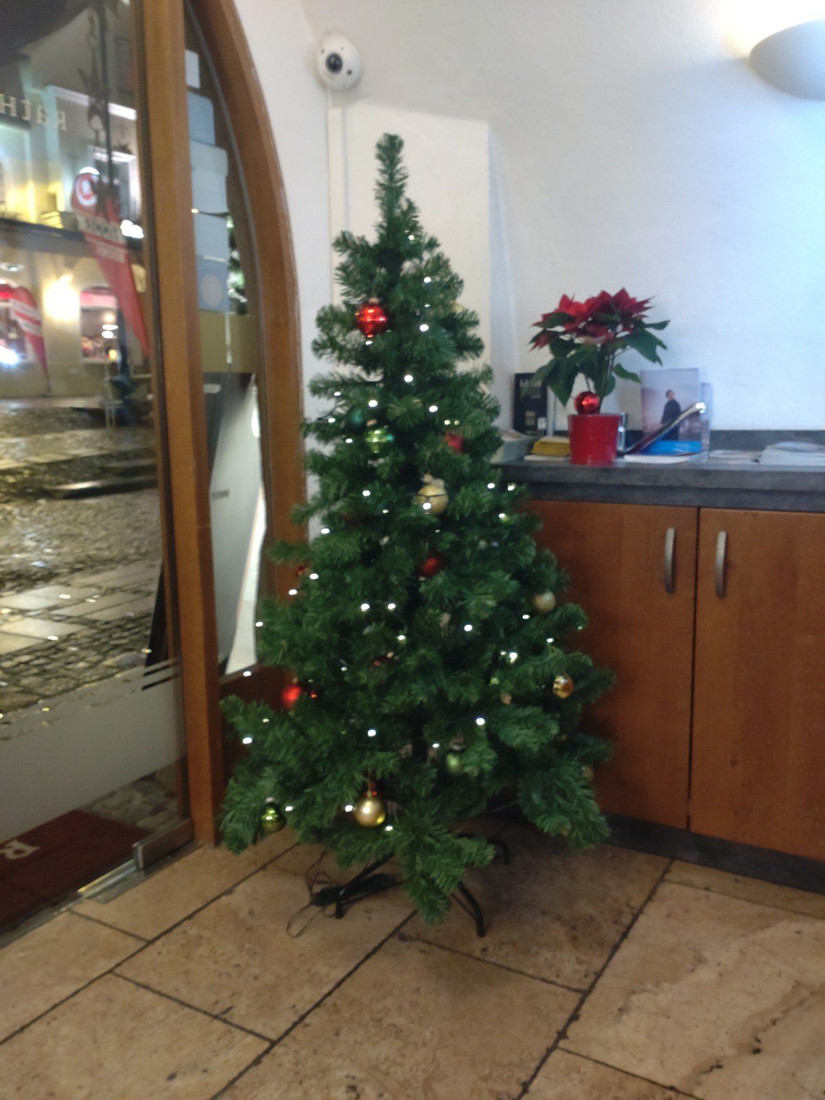 Ein Weihnachtsbaum ist mit Ornamenten und Lichtern geschmückt und steht vor einer Tür. Auf dem Tresen steht eine rote Weihnachtsstern in einem Topf.