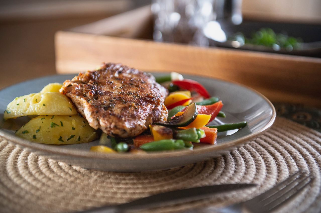Ein Teller mit gegrilltem Schweinekotelett, Kartoffelpüree und gemischtem Gemüse wie Karotten, Paprika und grünen Bohnen auf einem Tischset.
