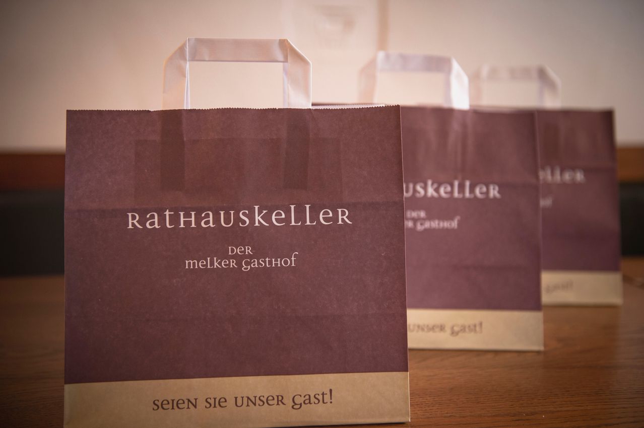 Ein brauner Papierbeutel mit 'Rathauskeller' und 'Der Melker Gasthof' darauf, auf einer Holzoberfläche platziert.