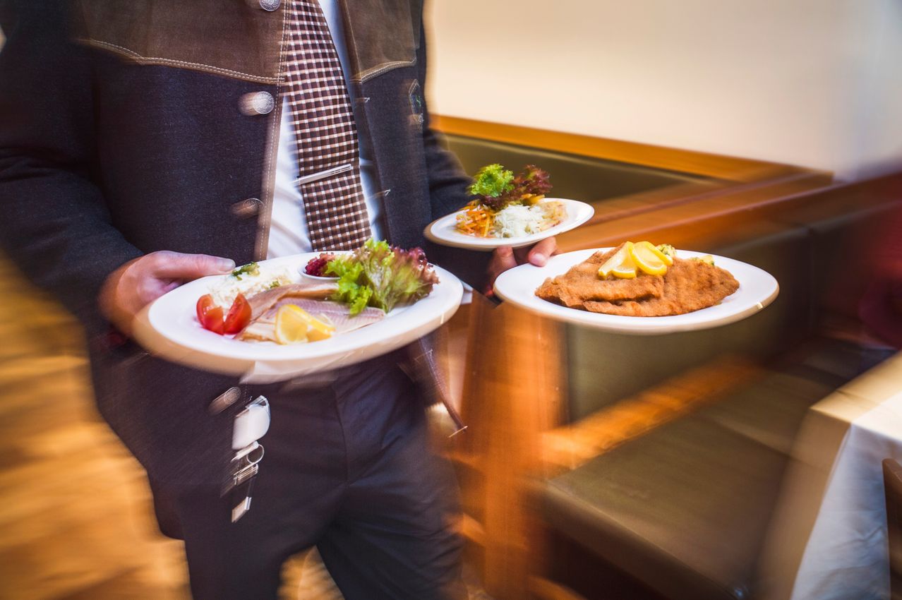 Ein Kellner trägt zwei Teller mit Essen. Ein Teller hat ein Fischgericht mit Tomaten und der andere ein gebratenes Gericht mit Zitronenscheiben.