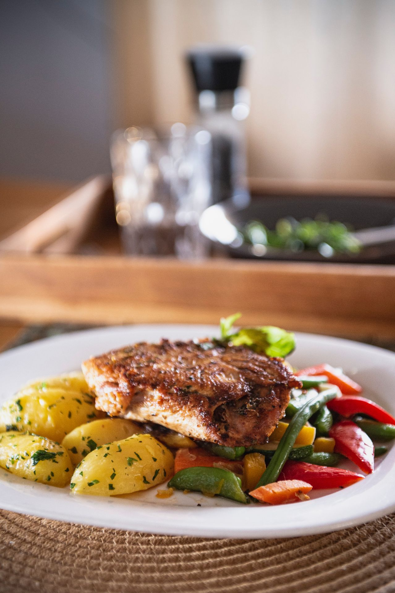 A white plate with grilled meat, yellow potatoes, green beans, and red bell peppers. A wooden tray and glass bottles are in the background.