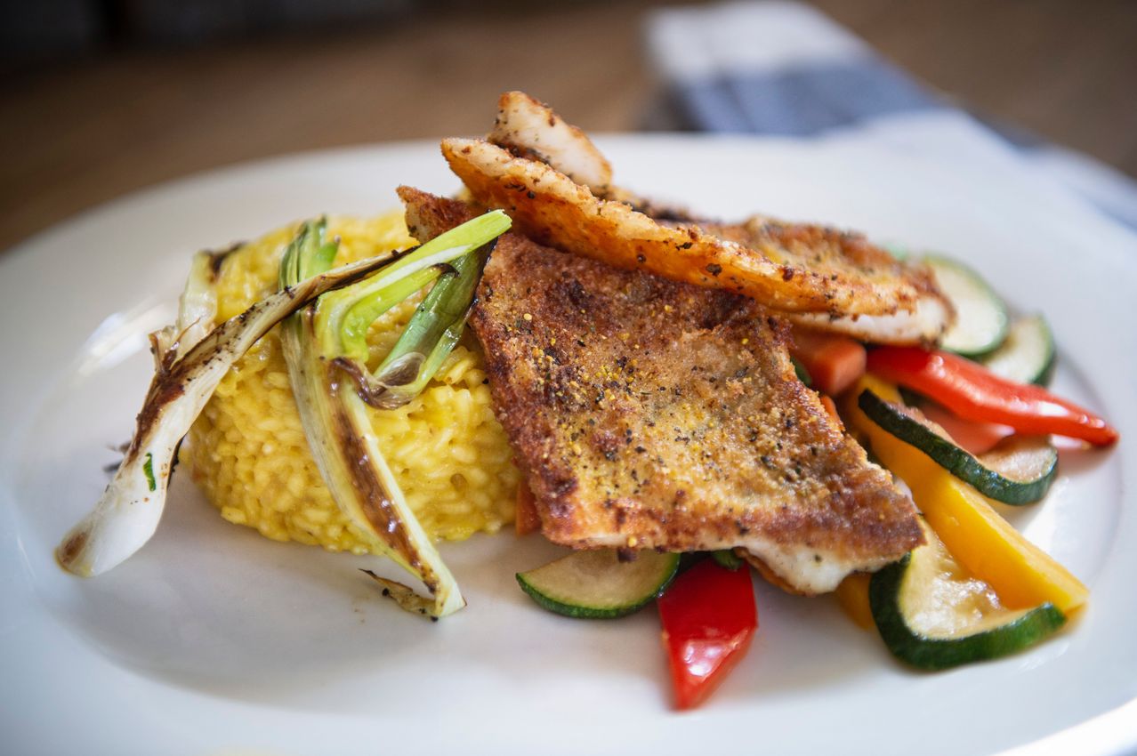 A white plate holds a fish fillet with seasonings, over rice and vegetables, including zucchini, bell peppers, and green onions.
