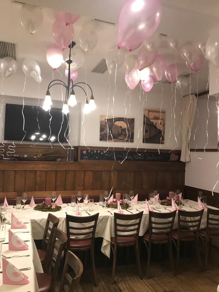 Ein Essbereich mit Tischen, die mit weißen Tüchern bedeckt sind, geschmückt mit rosa Servietten und Weingläsern. Herzförmige Luftballons hängen von der Decke. Zwei gerahmte Bilder hängen an der Wand.