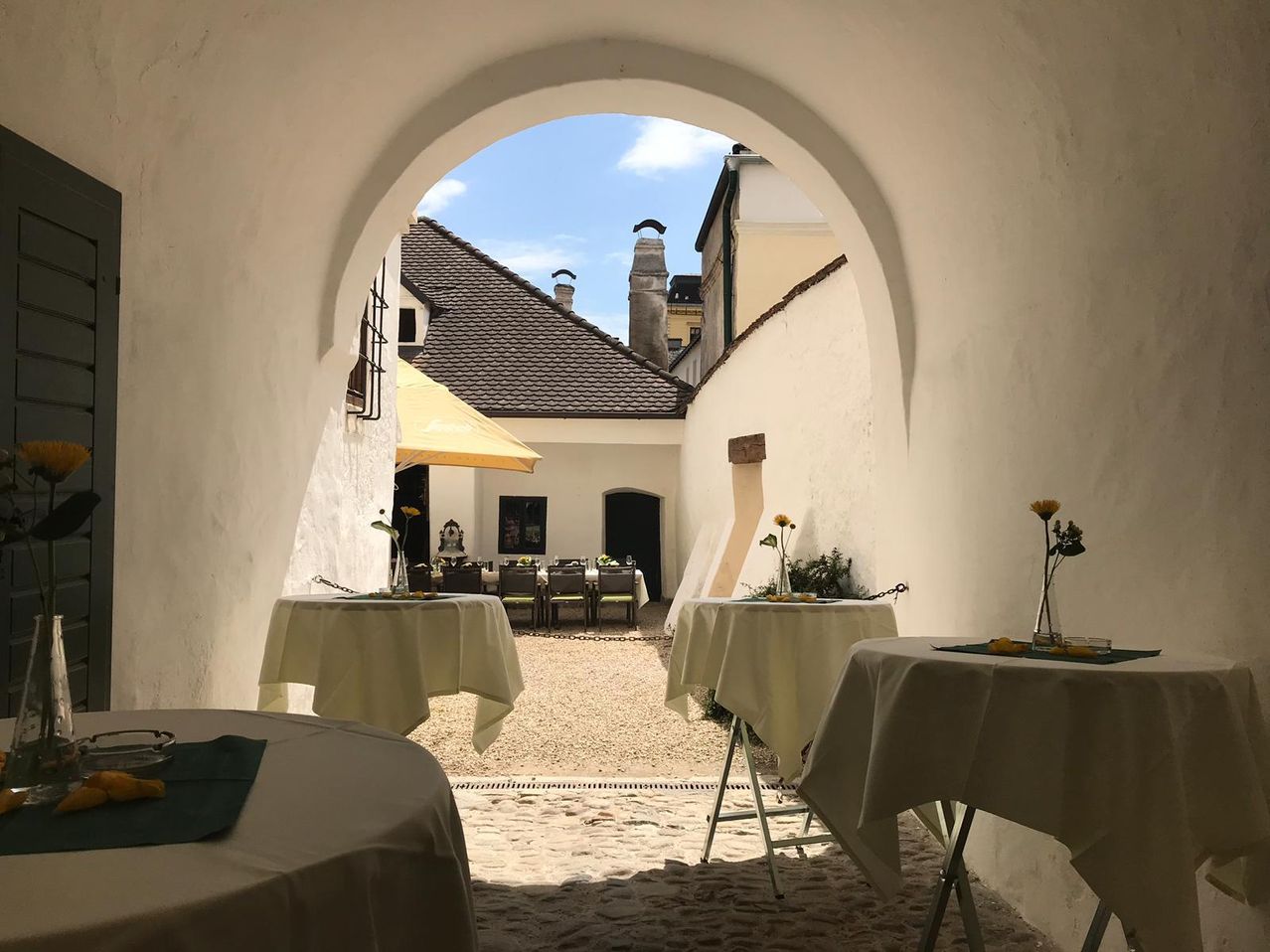 Ein Steinweg führt zu einem Bogen, der einen Innenhof mit drei Tischen unter einem gelben Regenschirm offenbart. Die Tische sind mit weißen Tüchern bedeckt und mit Blumen geschmückt. Hinter den Tischen befindet sich ein Essbereich mit Stühlen und einem großen Gebäude.
