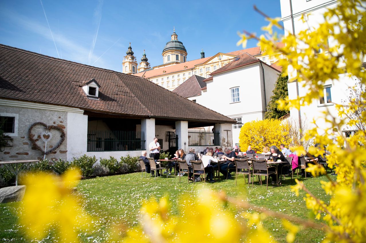 Several people are sitting at tables on a lawn in front of a building. A man stands near a table with a laptop. Behind him is a building with a brown roof. In the background is a historic building with towers and a dome.