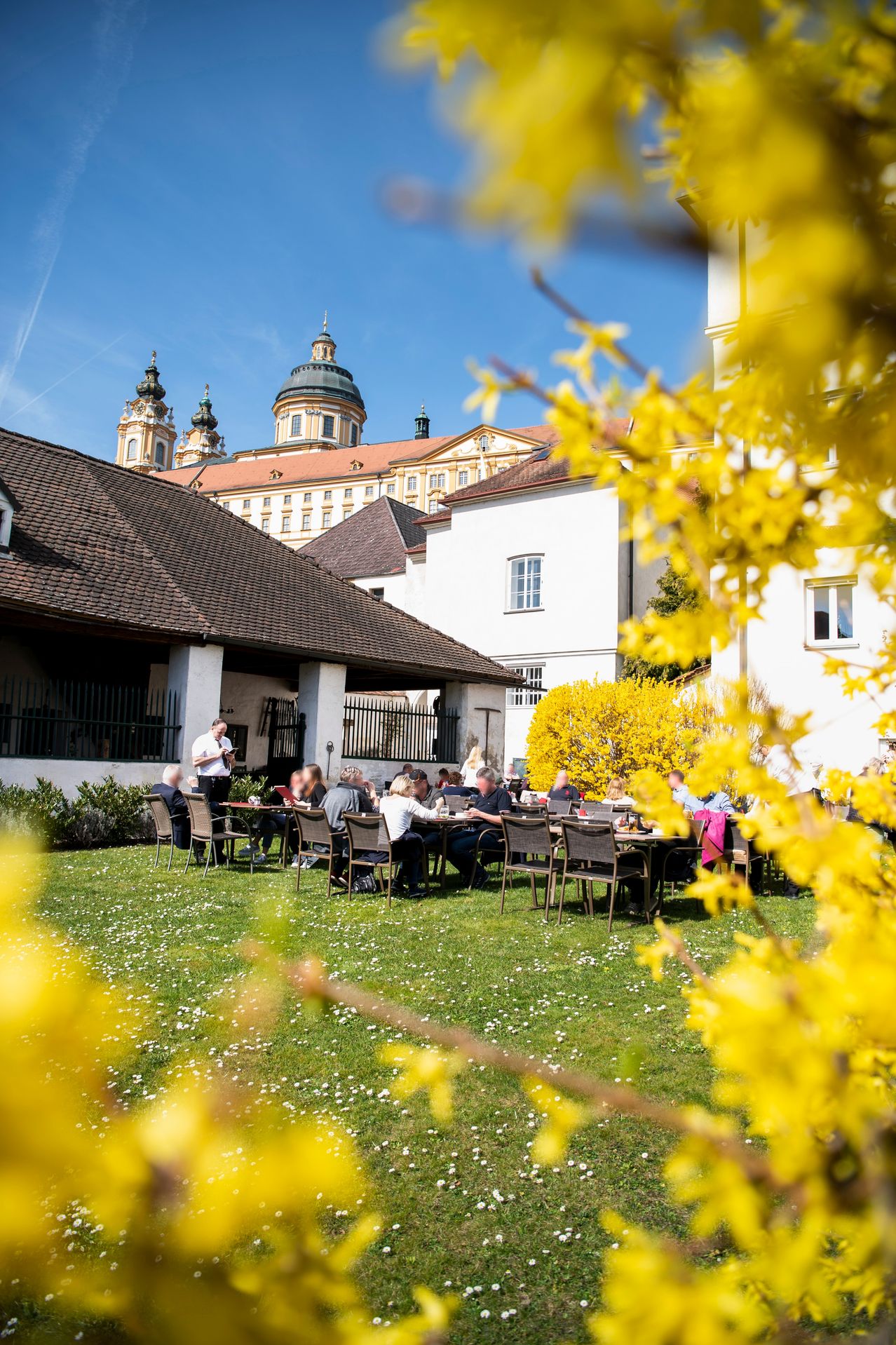 Eine Gruppe von Menschen isst im Freien in einem Restaurant mit Blick auf ein historisches Gebäude und gelbe Blumen im Vordergrund.