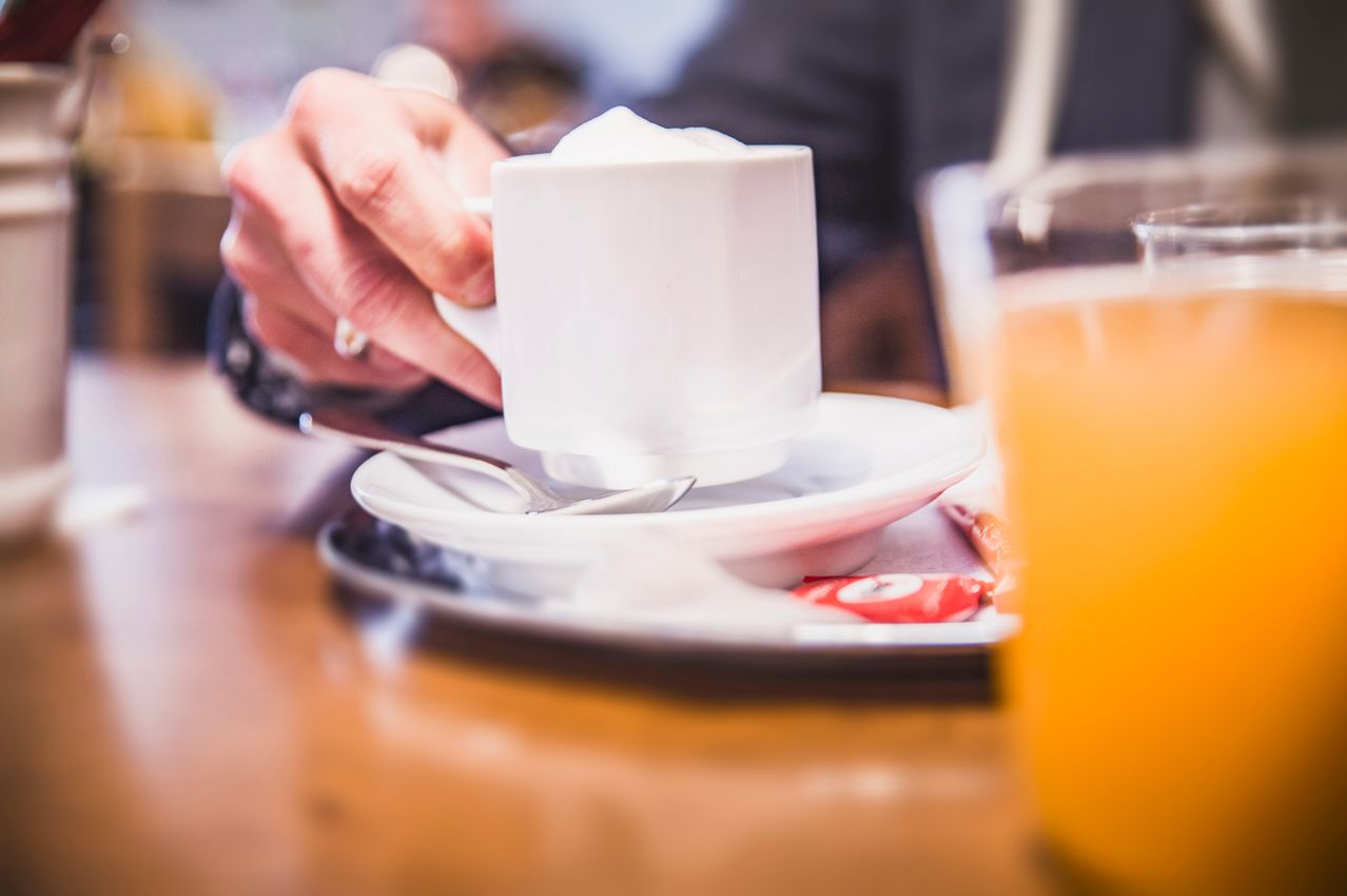 Eine Person hält eine weiße Kaffeetasse mit einem Löffel auf einer Untertasse, mit einem Glas Orangensaft in der Nähe.
