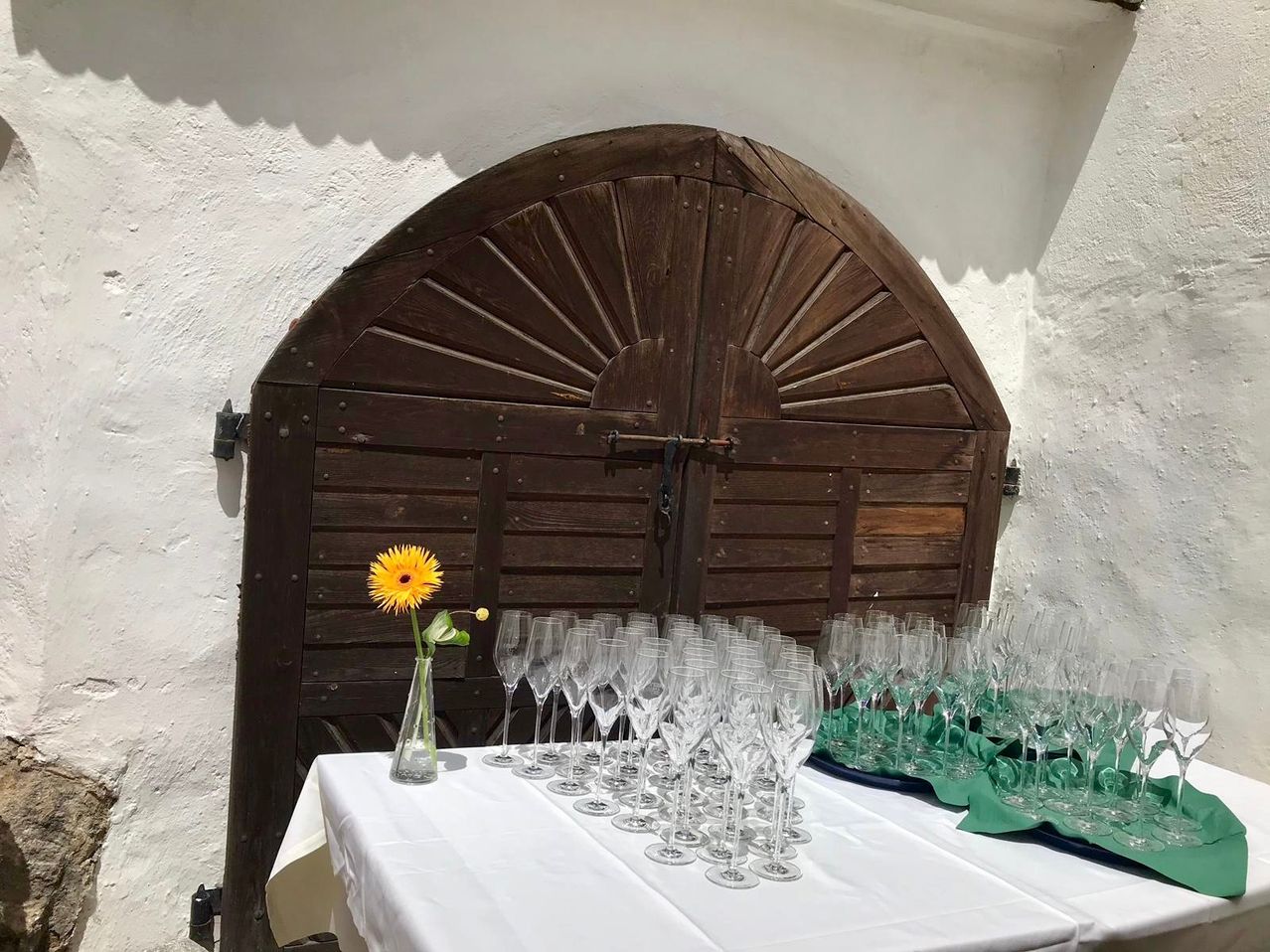 A table with a white tablecloth is set with many wine glasses and a yellow flower in a vase, with a wooden door in the background.