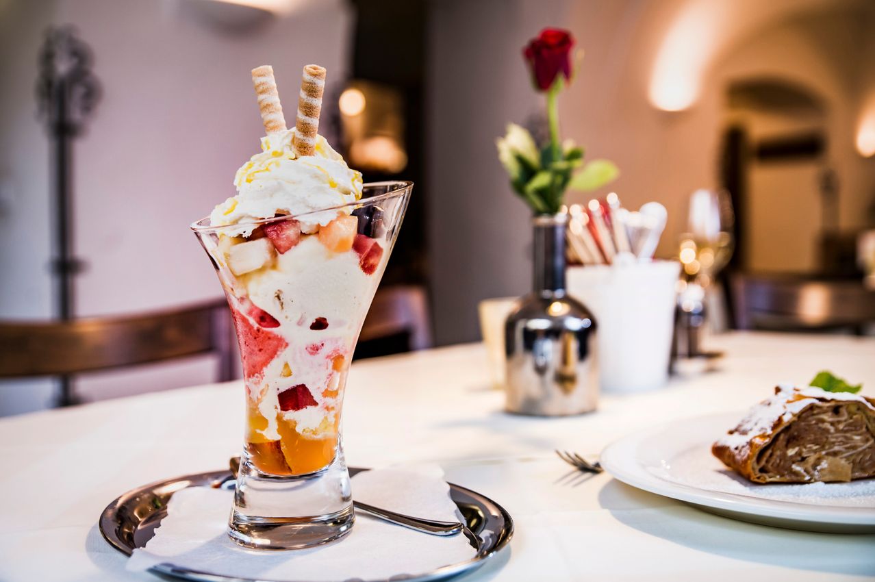 Ein Dessert mit Schichten aus Früchten, Eiscreme und Toppings auf einem Teller, mit einem Blumenstrauß im Hintergrund.