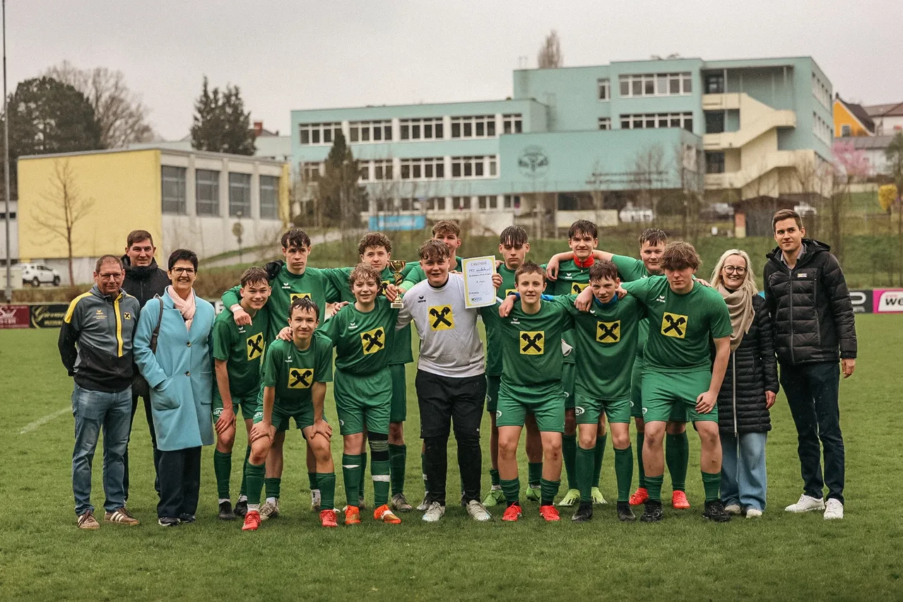 Eine Fußballmannschaft in grünen Trikots posiert für ein Foto auf einem Feld mit einem Pokal in der Hand. Dahinter steht ein Gebäude mit mehreren Fenstern.
