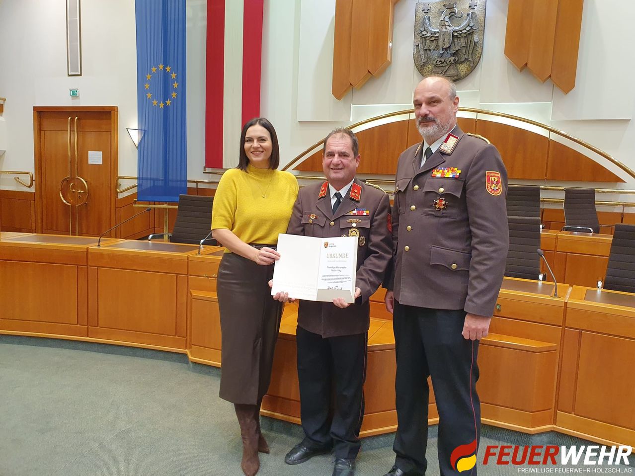 Eine Frau und zwei Männer in Uniformen stehen in einem Raum. Sie halten ein Zertifikat. Hinter ihnen sind ein Schreibtisch, Stühle und Flaggen zu sehen. Die Wand hat ein goldenes Design.