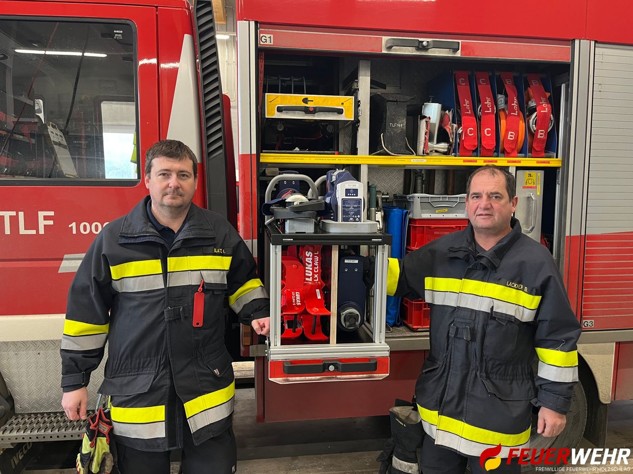 Zwei Feuerwehrleute stehen vor einem Feuerwehrwagen, einer hält einen Trolley mit medizinischer Ausrüstung. Die Fächer des Wagens sind mit verschiedenen Werkzeugen und Ausrüstungsgegenständen gefüllt.