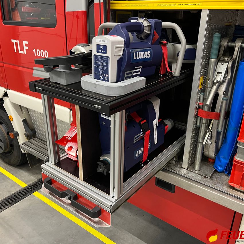 Ein offenes Fach eines Feuerwehrwagens mit einem blau-weißen LUKAS E3-Werkzeug auf einem schwarzen Regal und diversen Ausrüstungsgegenständen darum herum.