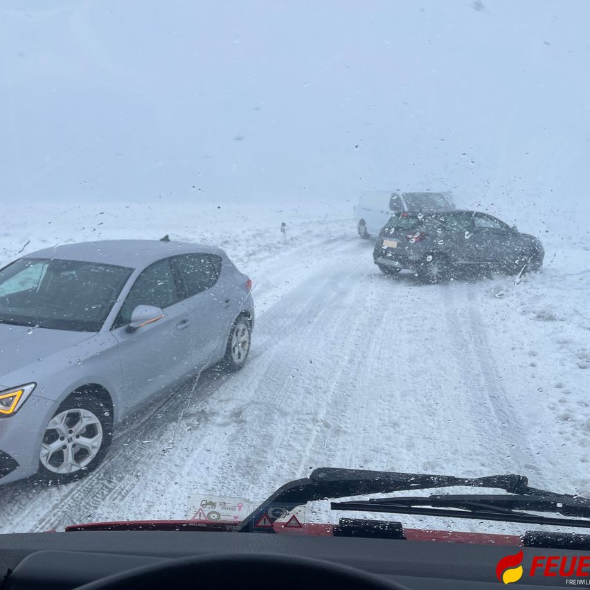 Eine verschneite Straße mit Autos, die in die gleiche Richtung fahren. Das vordere Auto hat seine Scheinwerfer eingeschaltet. Die Straße ist mit Schnee bedeckt.