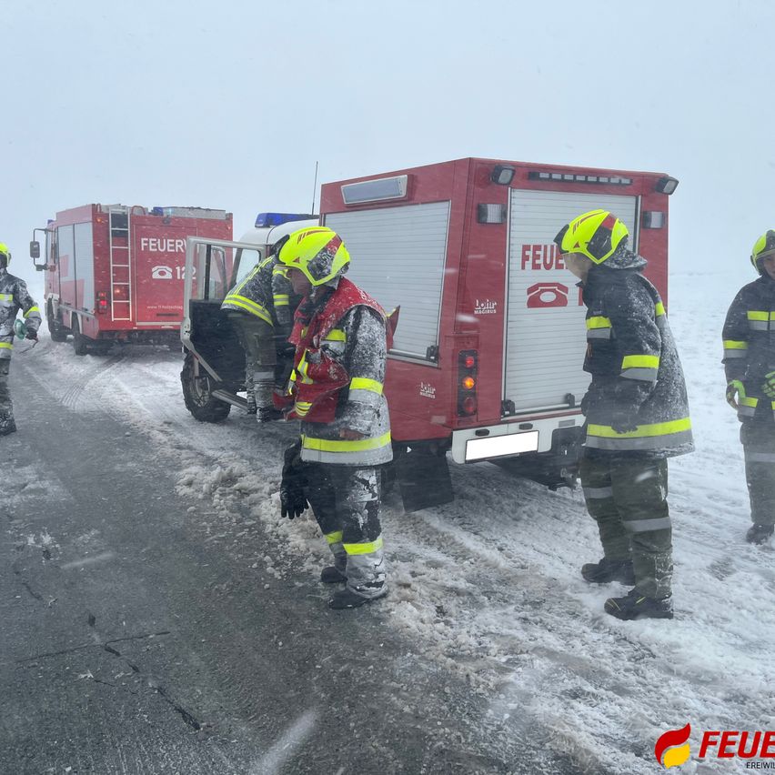 Feuerwehrleute in Schutzkleidung stehen auf einer verschneiten Straße. Zwei Feuerwehrwagen sind geparkt, einer mit offener Tür. Sie scheinen etwas zu besprechen.