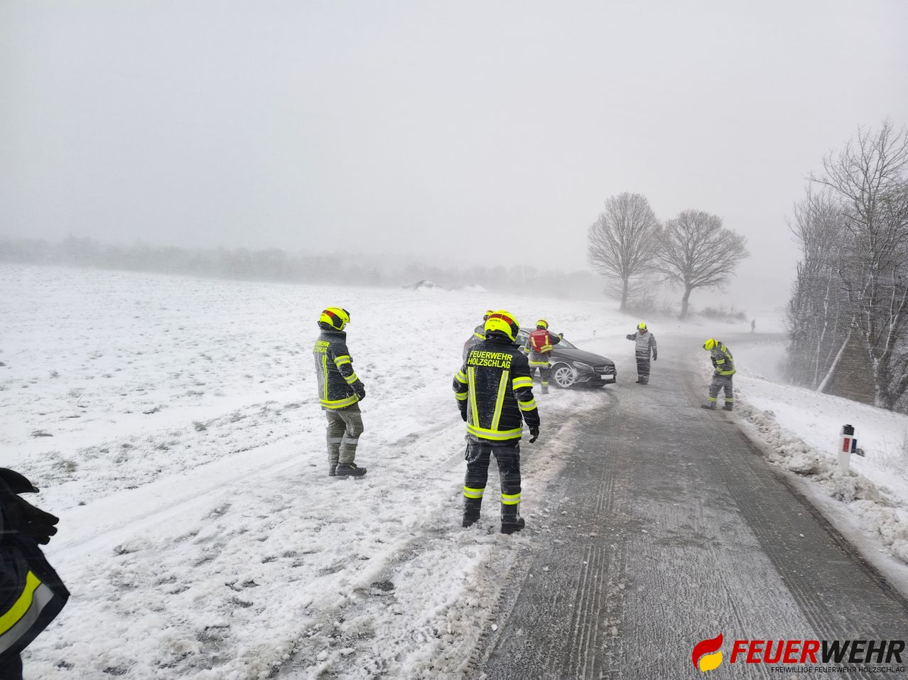 Eine Gruppe von Feuerwehrleuten in reflektierenden Kleidungsstücken steht auf einer verschneiten Straße, wo ein Auto geparkt ist. Sie reagieren wahrscheinlich auf einen Vorfall.