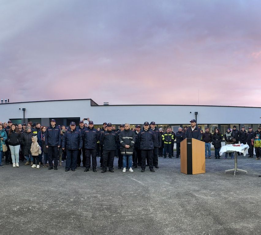 Eine Gruppe von Menschen steht vor einem Gebäude, einige in Uniformen, ein Mann spricht an einem Podium, ein Tisch hält Gegenstände.