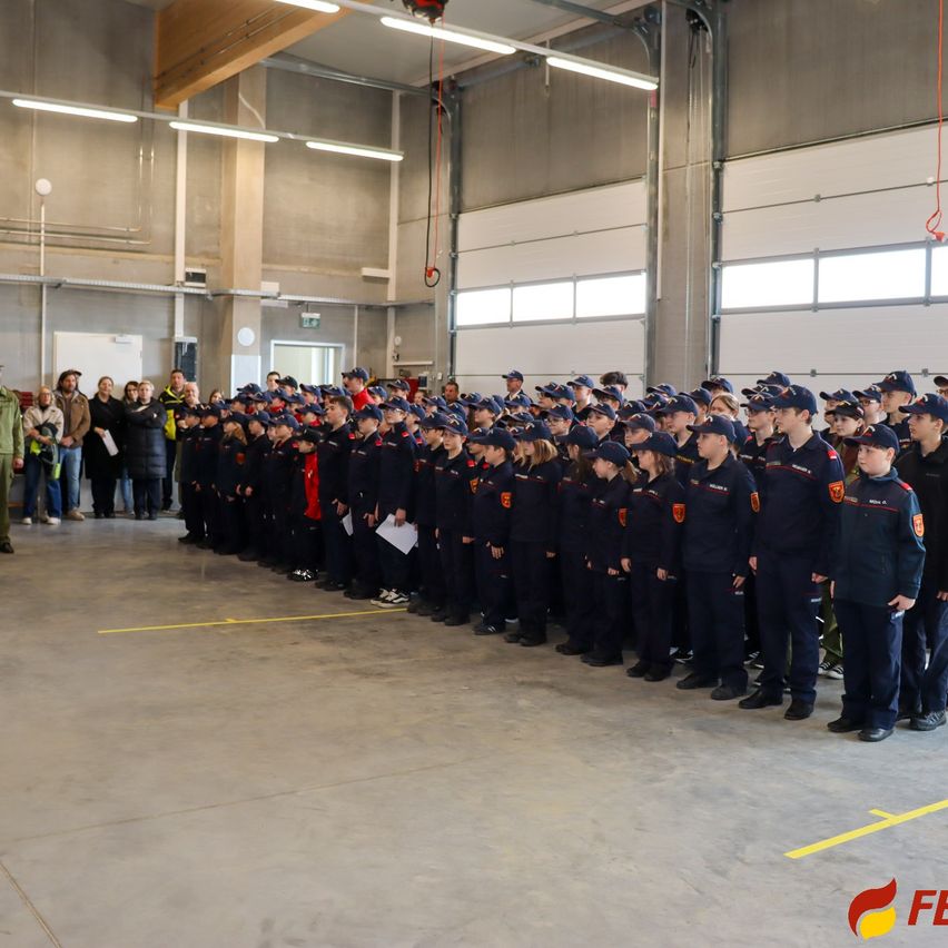Eine Gruppe uniformierter Personen steht in einer Reihe in einer Einrichtung, während Zuschauer von hinten zusehen. Die Uniformen haben auffällige Abzeichen.