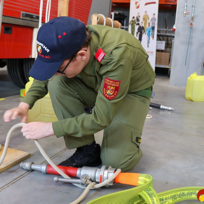 Ein Mann in einer grünen Uniform kniet auf dem Boden und justiert einen Schlauch, der mit einer rot-silbernen Feuerhydrante verbunden ist. Er trägt eine blaue Mütze und eine Brille. Ein roter und weißer Aufnäher befindet sich auf seiner Uniform. Ein gelber Eimer steht in der Nähe.