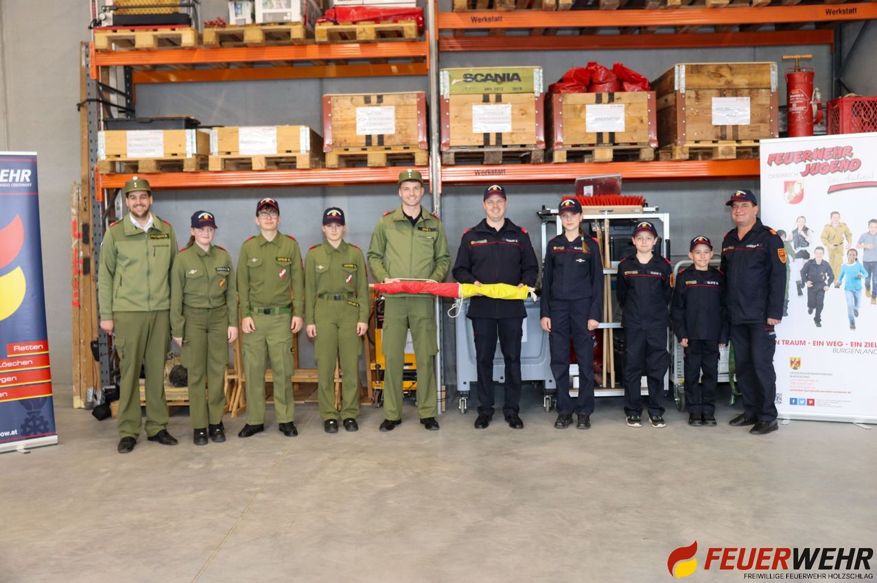 Eine Gruppe uniformierter Personen steht in einem Lagerhaus und hält eine rot-gelbe Flagge. Hinter ihnen sind Regale mit Kisten bestückt, darunter eine mit der Aufschrift 'Scania'.