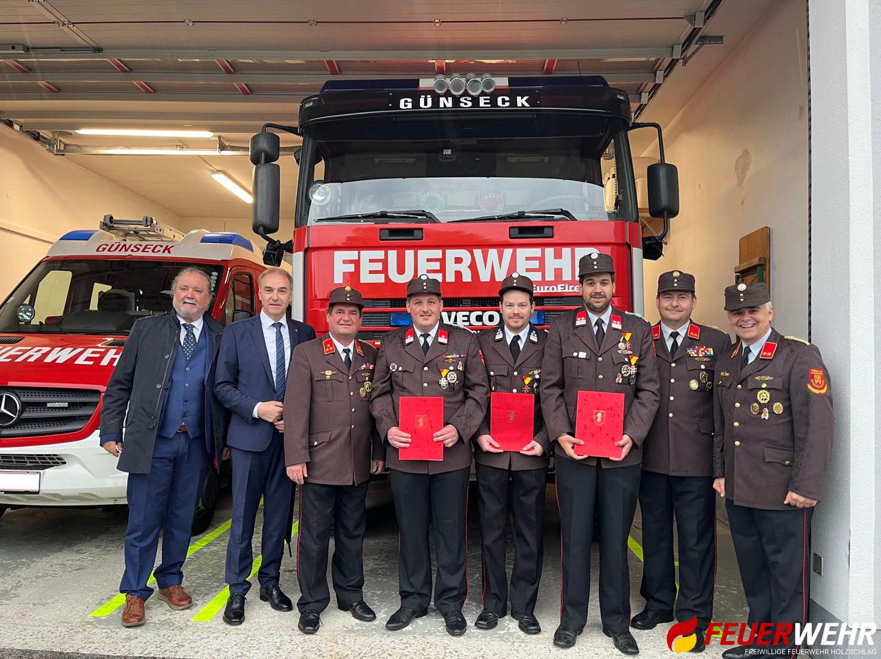Eine Gruppe von Feuerwehrleuten in Uniform steht vor einem roten LKW und hält Urkunden in den Händen. Zwei Männer in Anzügen stehen an den Seiten. Der LKW trägt die Aufschriften "FEUERWEHR" und "GUNSECK".