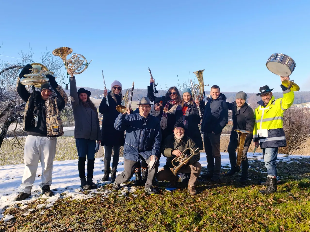 Eine Gruppe von Menschen in Winterkleidung posiert für ein Foto auf einem verschneiten Hügel und hält verschiedene Blechblasinstrumente in den Händen.