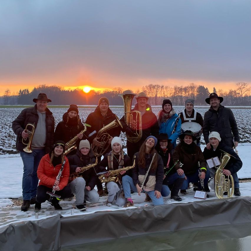 Eine Gruppe von Musikern in Winterkleidung posiert für ein Foto auf einem verschneiten Feld bei Sonnenuntergang. Sie halten verschiedene Instrumente wie Trompeten, Saxophone und Tuben.