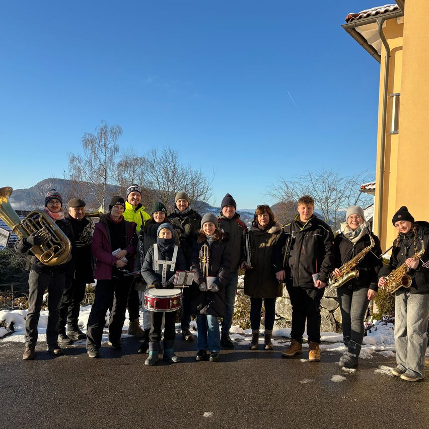 Eine Gruppe von Musikern posiert für ein Foto vor einem Gebäude mit schneebedecktem Hintergrund, in Winterkleidung und mit verschiedenen Musikinstrumenten.