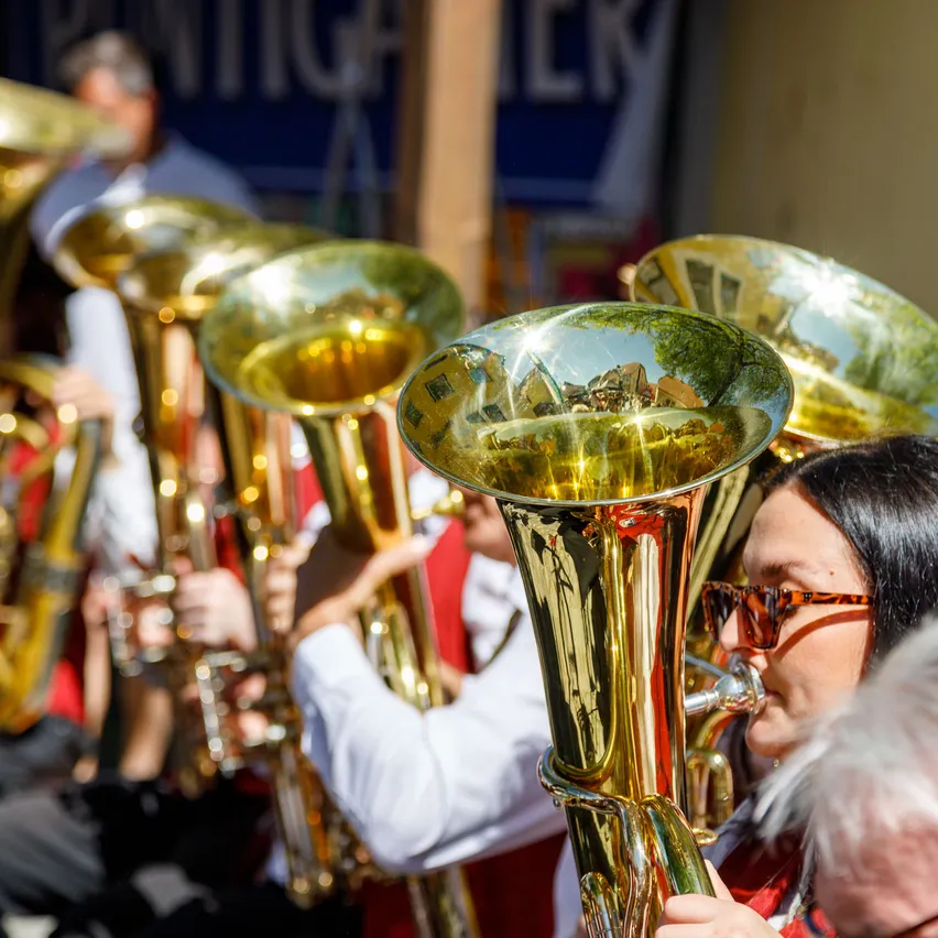 Eine Gruppe von Musikern in roten und weißen Uniformen spielt Blechblasinstrumente, möglicherweise bei einem Umzug. Die Instrumente sind golden und glänzend. Die Frau vorne spielt eine Tuba. Dahinter ist ein unscharfer Hintergrund.