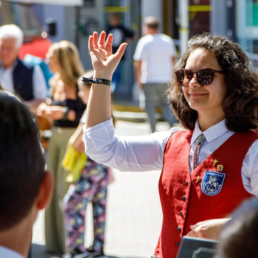 Eine Frau in einem roten Westchen winkt, während sie auf einer Straße zwischen anderen Menschen steht. Sie trägt eine Sonnenbrille und ein weißes Hemd.
