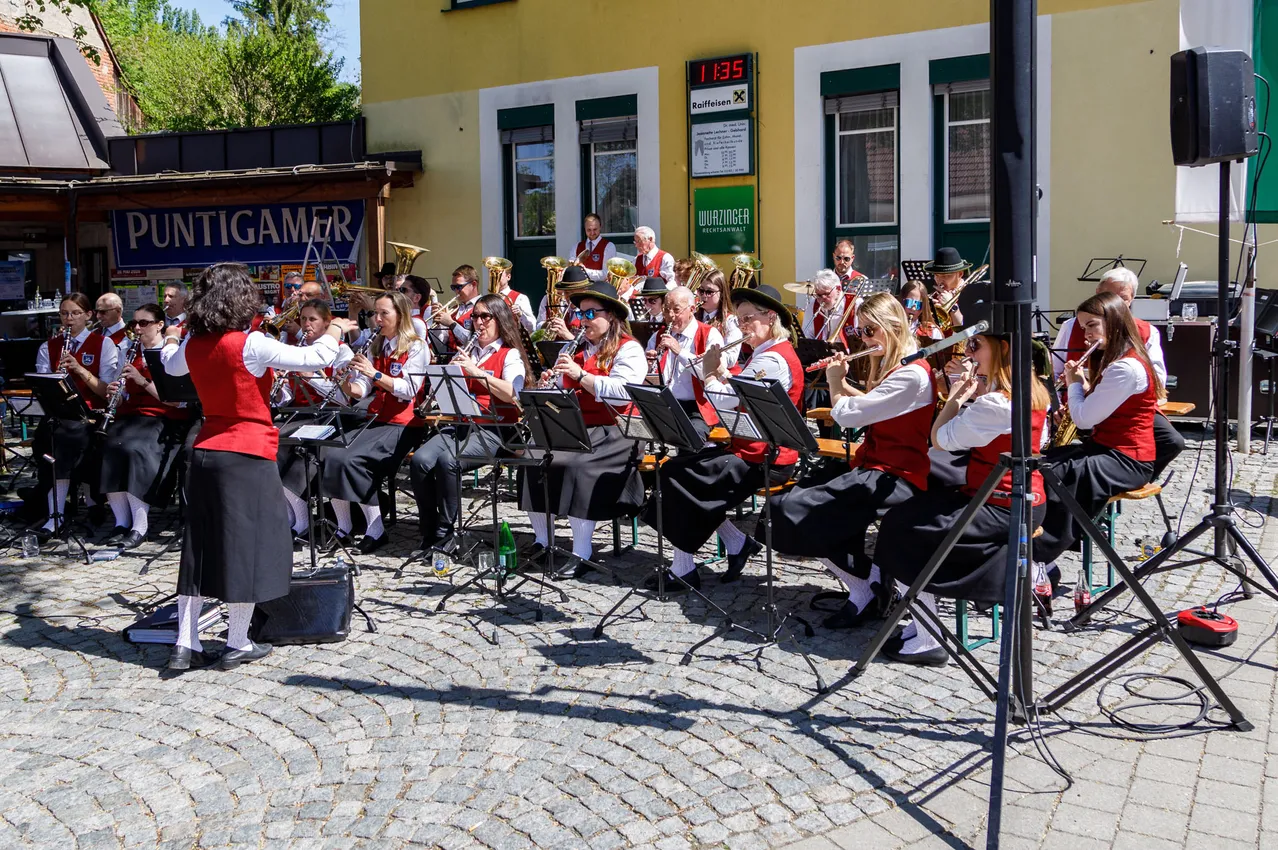 Eine Blechblaskapelle spielt auf einer Kopfsteinpflasterstraße, wobei die Musiker Instrumente wie Trompeten und Posaunen spielen. Das Wetter ist sonnig, und sie sitzen vor einem Gebäude.