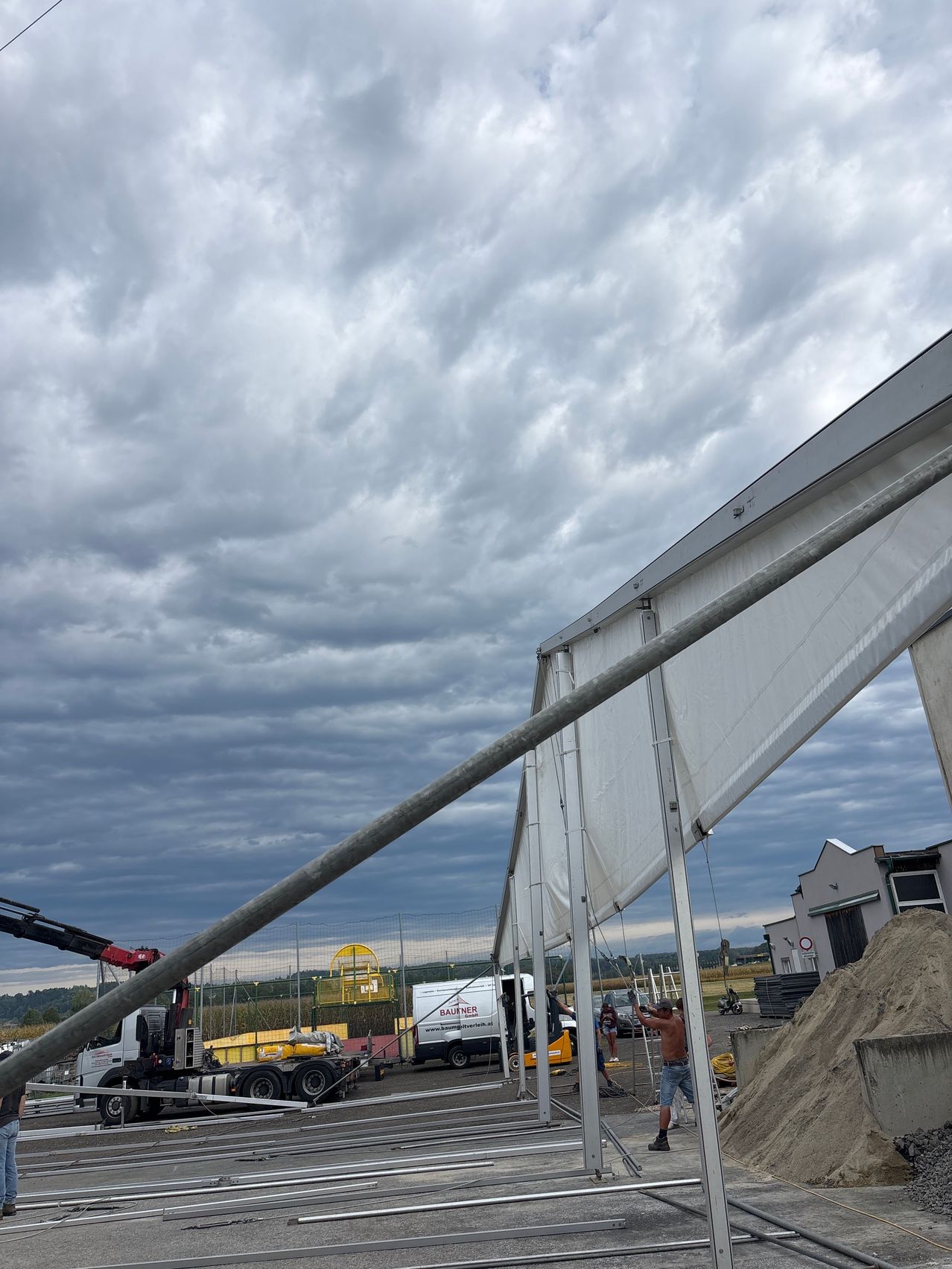 A construction site with a large tent being set up under a cloudy sky. A man is working on the tent while a truck is parked nearby.