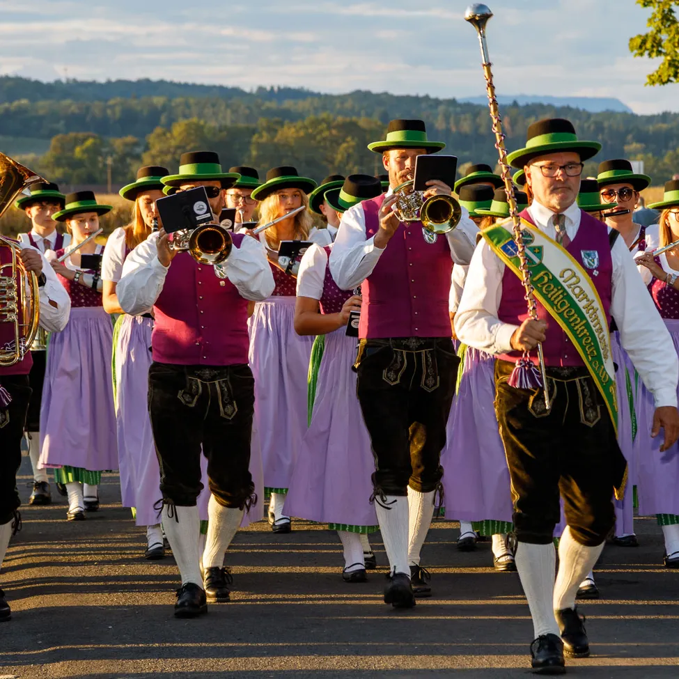 Eine Musikkapelle in traditioneller deutscher Kleidung tritt auf einer Straße auf, mit einer malerischen Kulisse im Hintergrund.