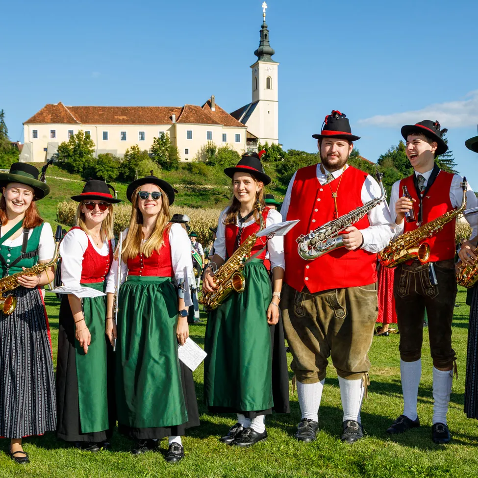 Eine Gruppe von Menschen, die in traditioneller deutscher Kleidung gekleidet sind, posiert für ein Foto, während sie Saxophon in einem Grasfeld halten.