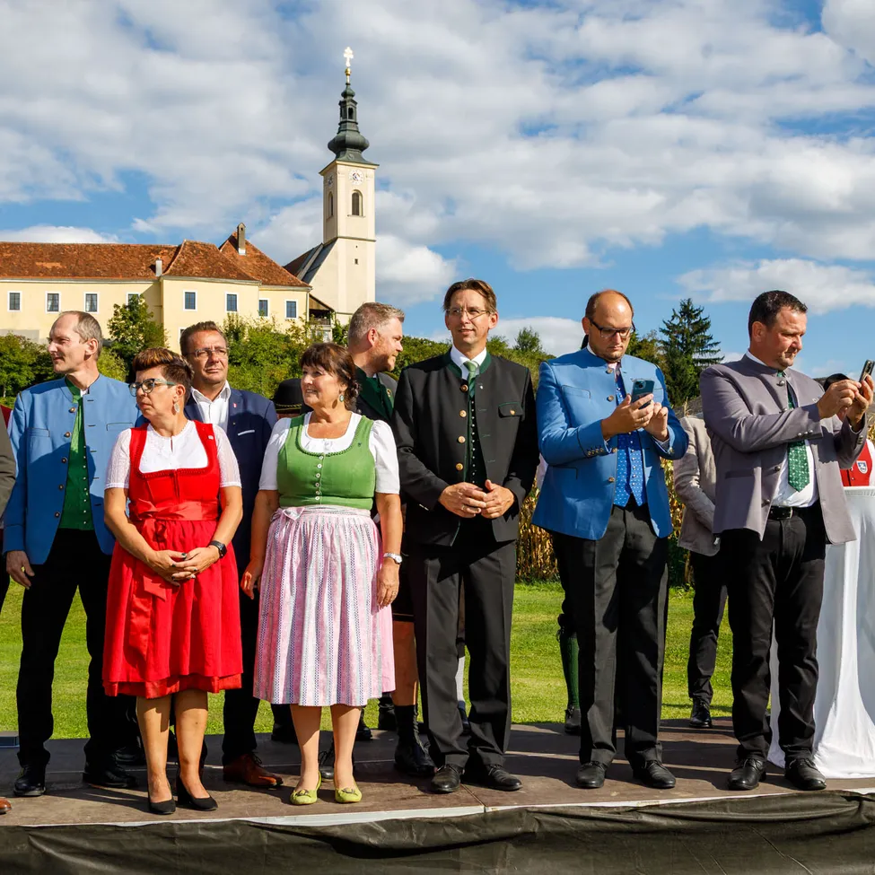 Eine Gruppe von Menschen steht auf einer Bühne vor einem Gebäude. Einige sind in traditioneller Kleidung gekleidet.