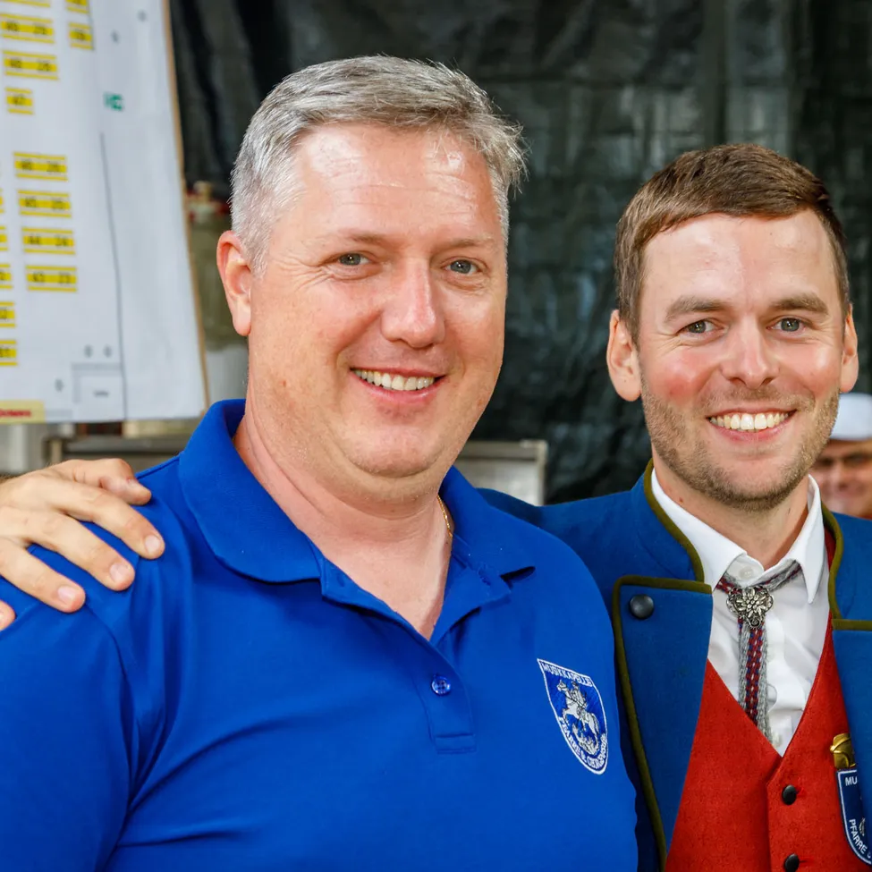 Zwei Männer lächeln und posieren für ein Foto. Der Mann auf der linken Seite trägt ein blaues Poloshirt und der Mann auf der rechten Seite trägt ein blaues und rotes Jackett mit einem weißen Innenshirt und einer Krawatte.
