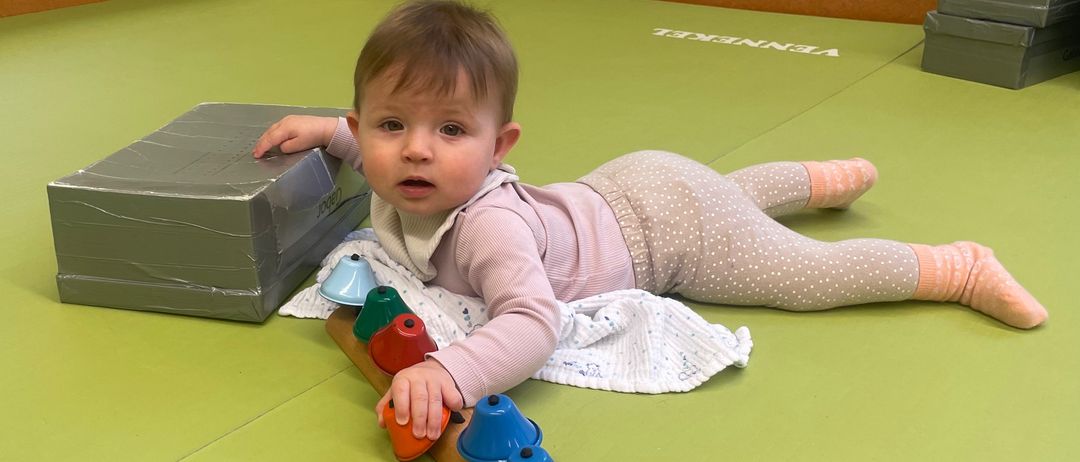 Ein Baby liegt auf einer grünen Matte und spielt mit einer Reihe bunter Glocken. Die Matte hat ein Logo in der Ecke.