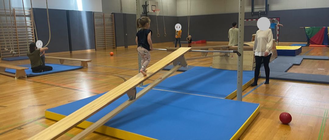 Ein kleines Mädchen balanciert auf einem Holzbalken über einer blauen Matte in einer Turnhalle, während andere in der Nähe sind. Es sind mehrere andere Geräte der Turnhalle sichtbar.