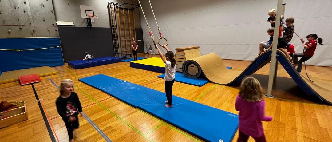 Eine Gruppe von Kindern spielt in einer Turnhalle. Ein Kind steht auf einer blauen Matte und hält einen Ring. Ein anderes Kind sitzt auf einer Rutsche. Einige Kinder spielen an der Kletterwand.