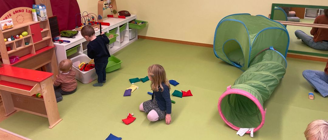 Kinder spielen in einem Raum mit grünem Boden, Spielzeug und einem Zelt. Ein Junge schaut sich Spielzeug an, ein Mädchen sitzt mit bunten Bausteinen.