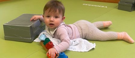 Ein Baby liegt auf einem grünen Boden und spielt mit einem hölzernen Xylophon mit bunten Glocken. Das Baby trägt ein rosa Outfit und hält das Xylophon. Ein Handtuch ist neben dem Baby auf dem Boden ausgebreitet.