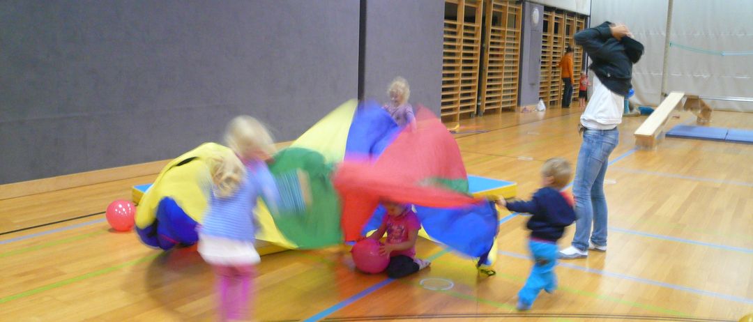 Mehrere Kinder spielen mit einem großen bunten Fallschirm in einer Turnhalle. Einige rennen darum herum, während andere darin sitzen. Eine Person beobachtet von der Seite.
