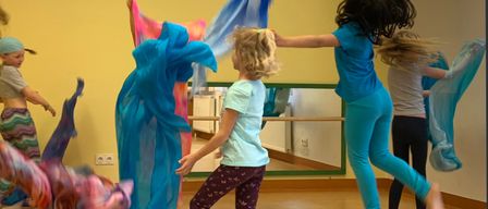 Mehrere Kinder in einem Studio mit farbenfrohen Tüchern tanzen, wobei ein Kind im Vordergrund springt und zwei weitere hinter ihr sind.