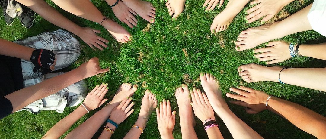 Eine Gruppe von Menschen sitzt im Kreis auf Gras, ihre Füße berühren sich und bilden ein Kreismuster. Sie tragen Armbänder und haben grünen Nagellack.